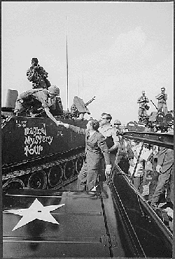 Nixon standing beside tank in Vietnam.  Soldiers on tank reach down to shake his hand